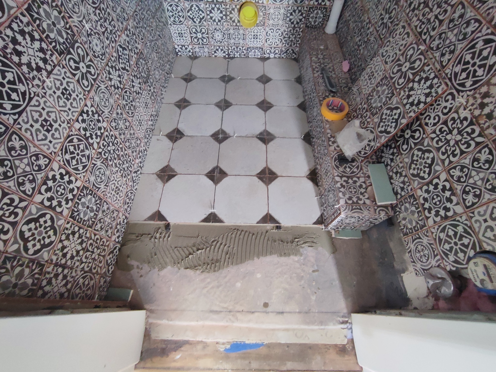 Układanie wzorzystych płytek podłogowych i ściennych w łazience, widoczna zaprawa klejowa na podłodze, plastikowe krzyżyki dystansowe i narzędzia glazurnicze.