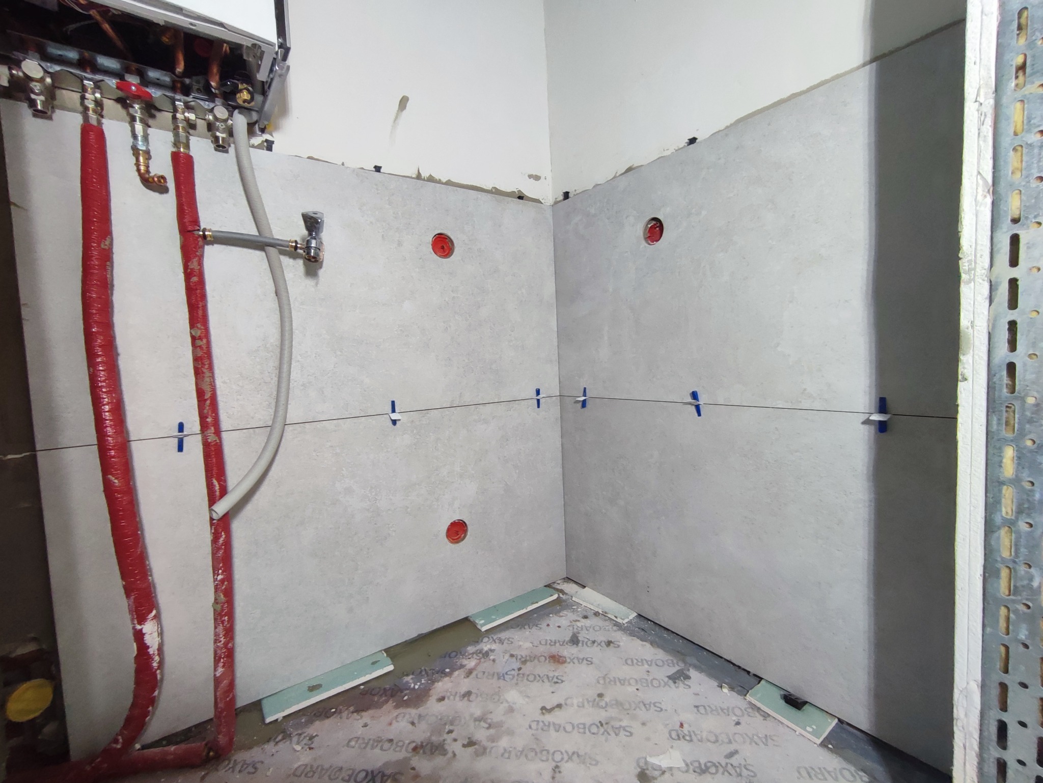 Ściany w trakcie układania płytek, widoczne przewody instalacji wodnej z czerwoną izolacją, poziomica laserowa i krzyżyki dystansowe do płytek.