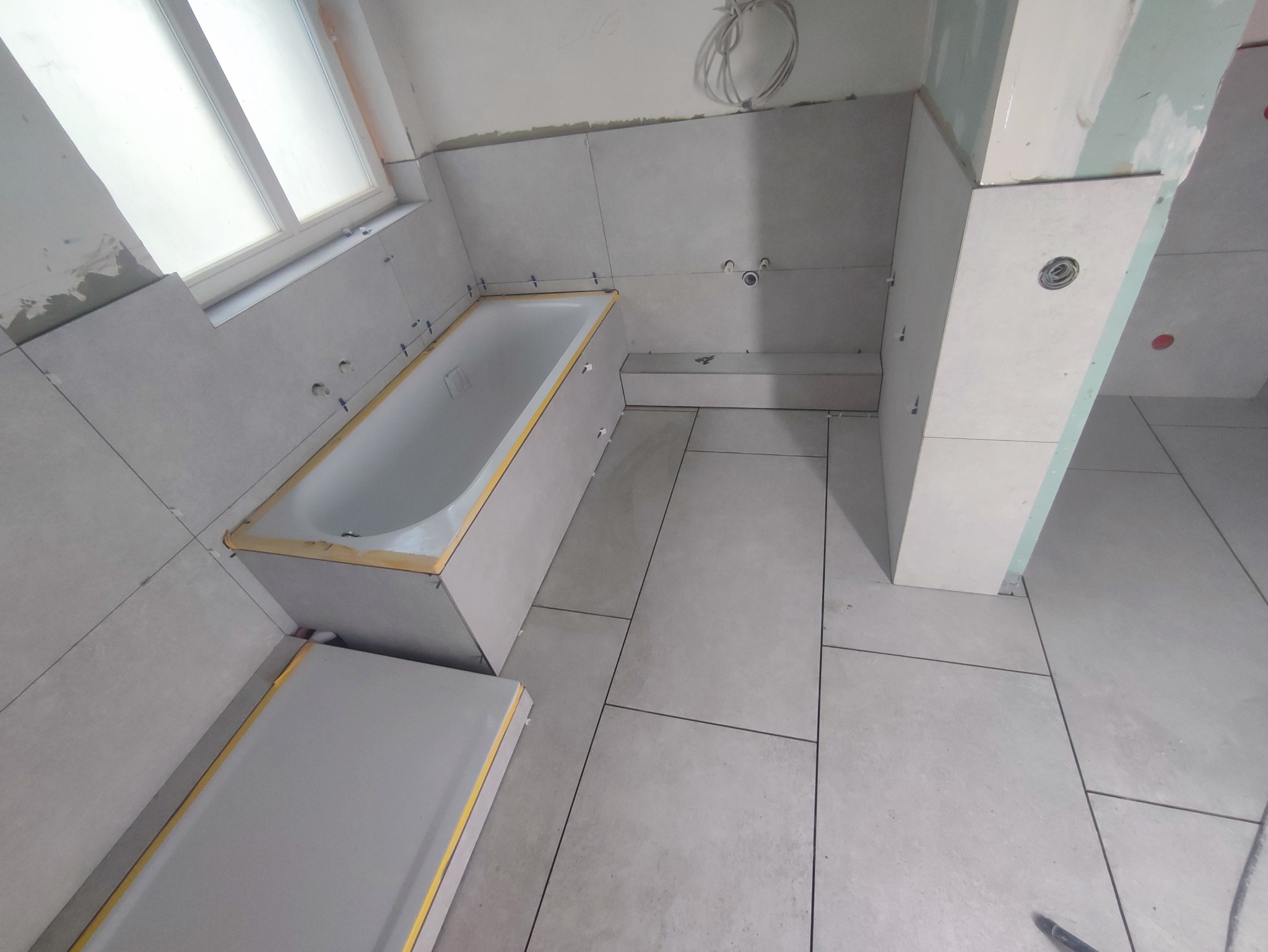 Łazienka w trakcie remontu: montaż szarych płytek na ścianach i podłodze, obudowa wanny zabezpieczona taśmą malarską, widoczne krzyżyki glazurnicze.
