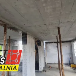 Wnętrze surowego budynku w trakcie budowy, ściany z białych bloczków, betonowy strop podparty drewnianymi stemplami, widoczne okna i drzwi w stanie surowym, w tle logo firmy budowlanej 'Perfect...