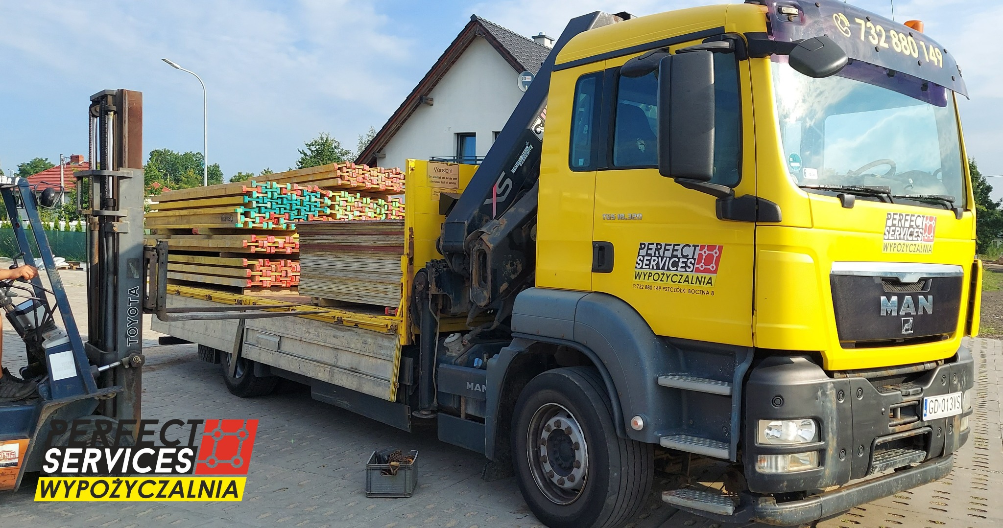 Żółta ciężarówka MAN z dźwigiem załadowana drewnianymi belkami, obok wózek widłowy Toyota przygotowuje się do podniesienia ładunku. Widoczne logo firmy Perfect Services Wyposażalnia.