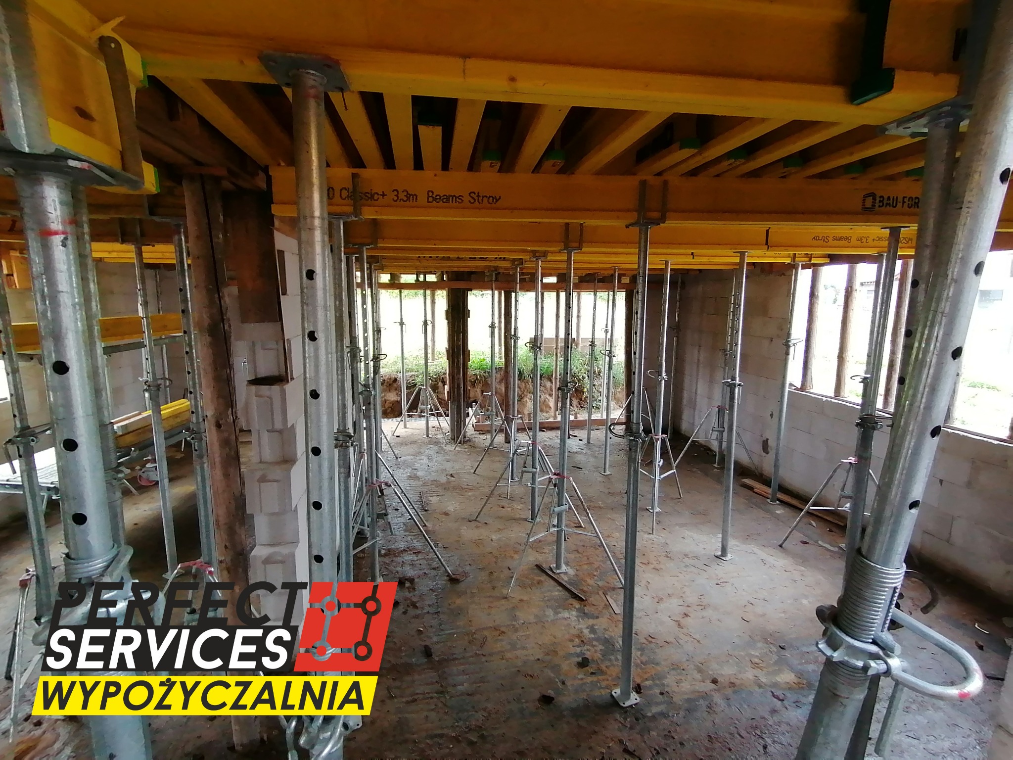 Wnętrze surowego budynku ze stemplami budowlanymi podtrzymującymi drewniany strop, widoczny logotyp wypożyczalni sprzętu budowlanego w lewym dolnym rogu.