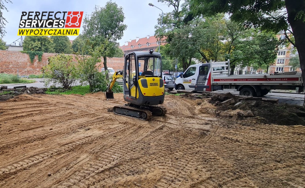 Żółta mini koparka Wacker Neuson na placu budowy, widoczne ślady gąsienic na piasku, w tle samochód dostawczy z napisem 'Rusztowania wynajem sprzedaż' oraz ceglany mur.