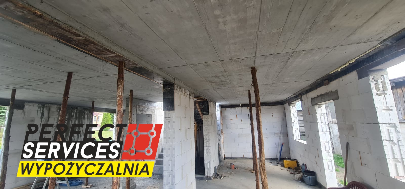 Wnętrze surowego budynku w trakcie budowy, ściany z białych bloczków, betonowy strop podparty drewnianymi stemplami, widoczne okna i drzwi w stanie surowym, w tle logo firmy budowlanej 'Perfect...