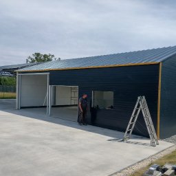 ORNARE sp. z o.o. - Montaż hali z blachy trapezowej: widok z zewnątrz na otwartą bramę segmentową, drabina oparta o ścianę, pracownik w tle. Nowoczesna konstrukcja z elementami wykończeniowymi.