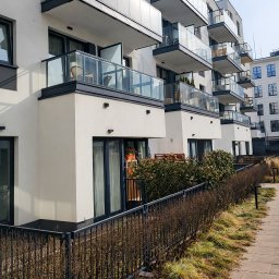 ORNARE sp. z o.o. - Nowoczesny budynek z balkonami z balustradami szklanymi, widok z dołu, z niskim ogrodzeniem metalowym wzdłuż chodnika. Jasne, naturalne światło.