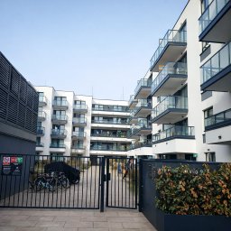 ORNARE sp. z o.o. - Otwarta, czarna brama wjazdowa z pionowych prętów, prowadząca na dziedziniec nowoczesnego osiedla z białymi budynkami i szklanymi balustradami balkonów.