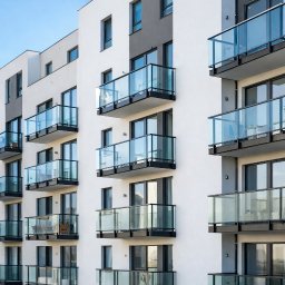 ORNARE sp. z o.o. - Elewacja budynku z balkonami z balustradami szklanymi i czarnymi ramami, widok z dołu, błękitne niebo w tle. Na balkonach widoczne rośliny w donicach.