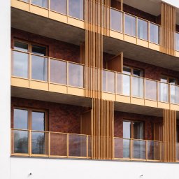 ORNARE sp. z o.o. - Nowoczesny budynek z balkonami z balustradami szklanymi i drewnianymi elementami dekoracyjnymi. Ujęcie z dołu, podkreślające geometrię i detale wykończenia.