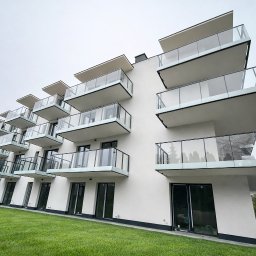 ORNARE sp. z o.o. - Nowoczesny budynek z białymi balkonami i szklanymi balustradami, widok z dołu na tle zielonego trawnika i pochmurnego nieba. Minimalistyczna architektura.