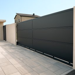 ORNARE sp. z o.o. - Nowoczesna, przesuwna brama wjazdowa w antracytowym kolorze, z subtelnymi podziałami poziomymi, na tle jasnej kostki brukowej i fragmentu domu. Minimalistyczny design.