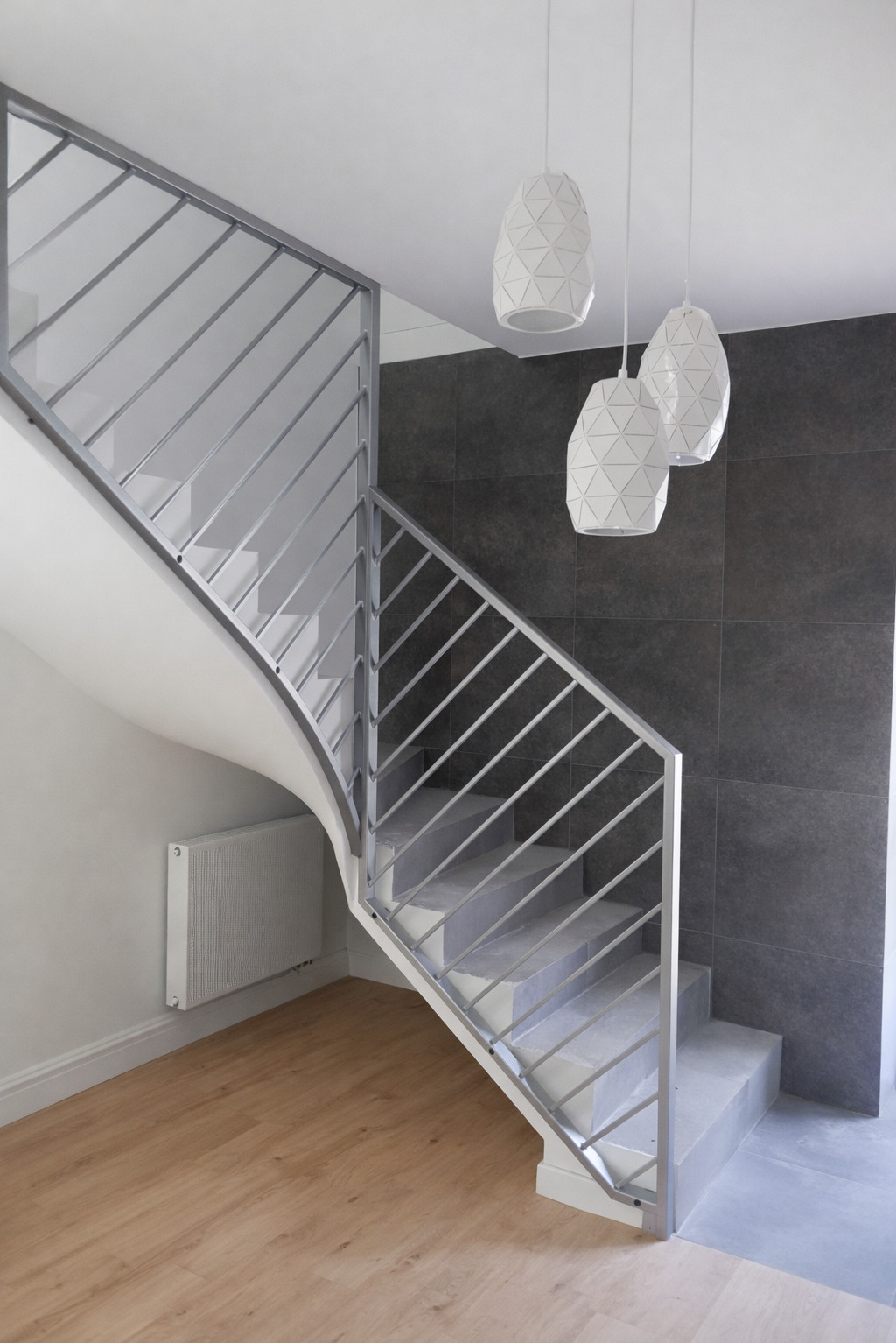 Nowoczesne schody z metalową balustradą o diagonalnym wzorze, oświetlone trzema lampami wiszącymi. Szare płytki na ścianie i drewniana podłoga dodają elegancji.
