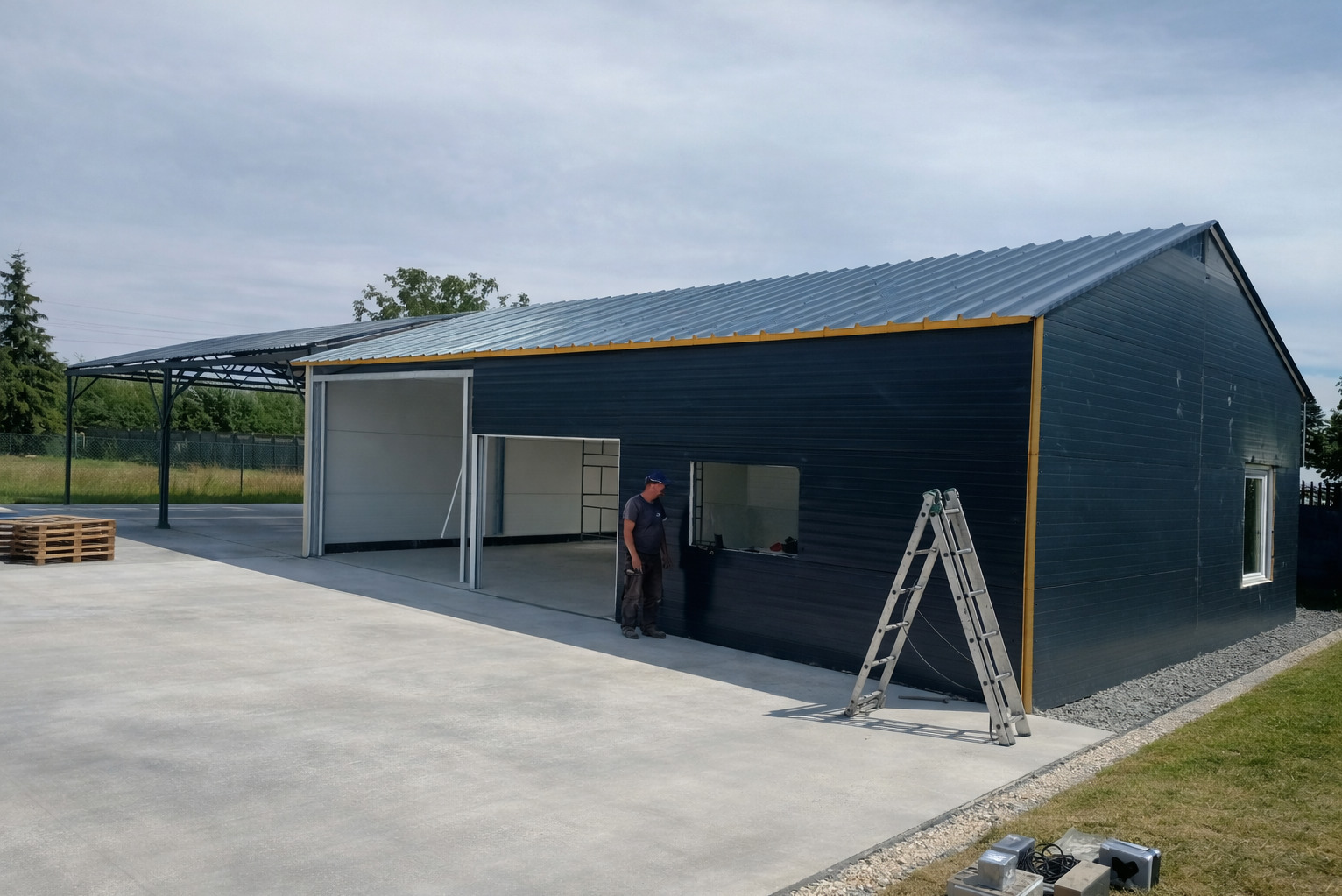 Montaż hali z blachy trapezowej: widok z zewnątrz na otwartą bramę segmentową, drabina oparta o ścianę, pracownik w tle. Nowoczesna konstrukcja z elementami wykończeniowymi.