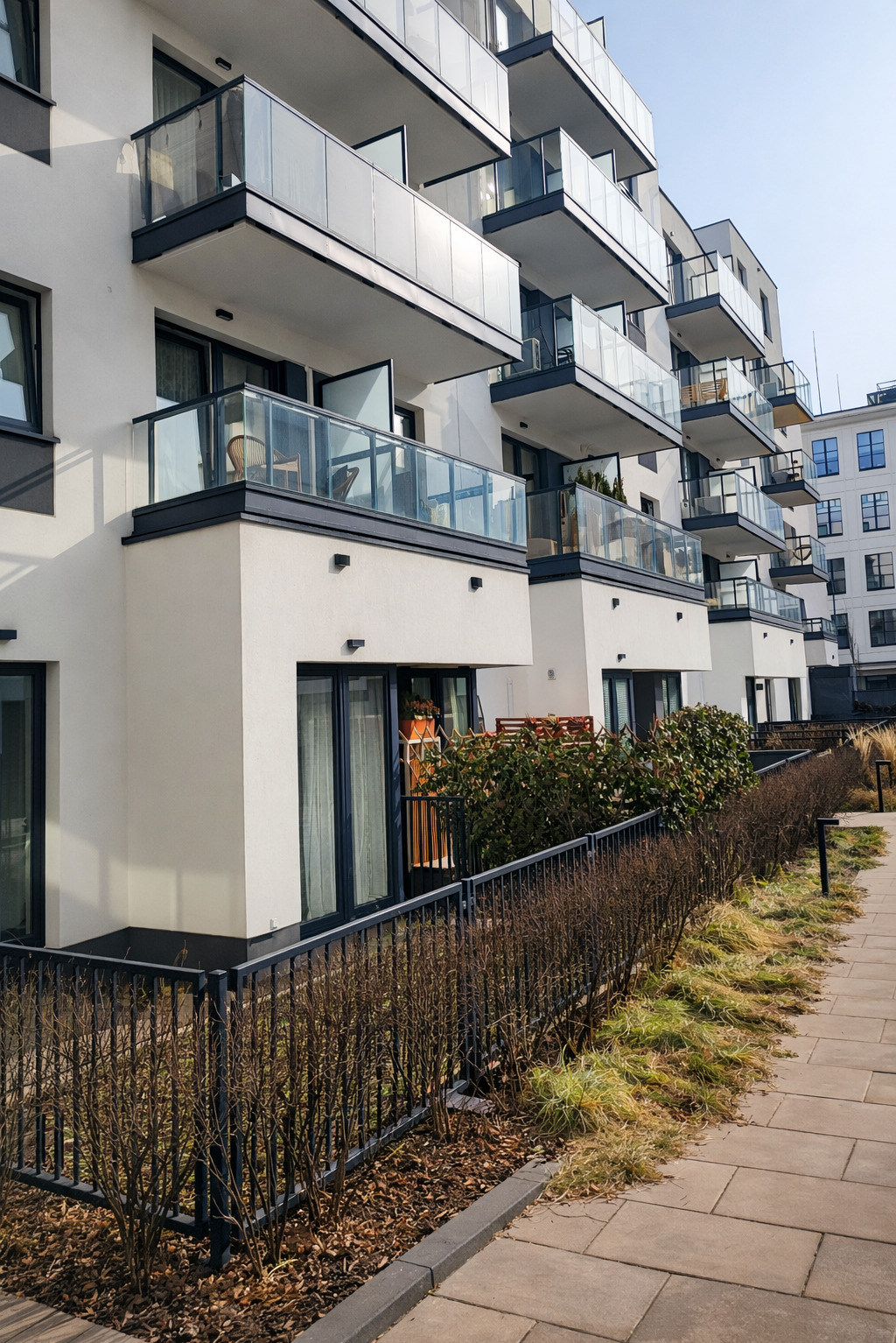 Nowoczesny budynek z balkonami z balustradami szklanymi, widok z dołu, z niskim ogrodzeniem metalowym wzdłuż chodnika. Jasne, naturalne światło.