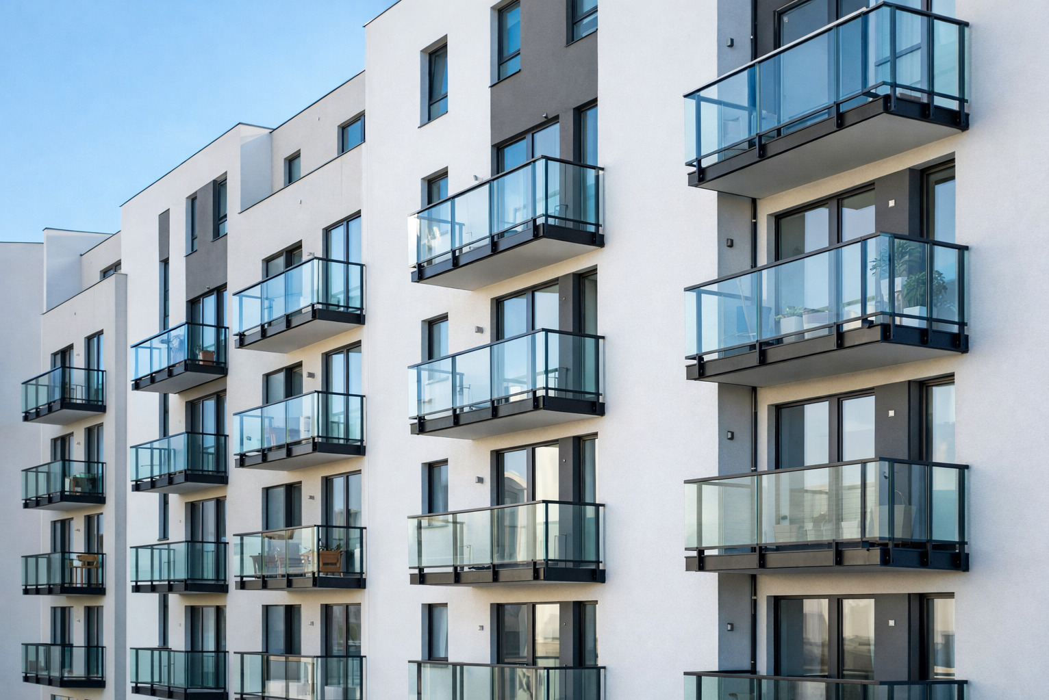 Elewacja budynku z balkonami z balustradami szklanymi i czarnymi ramami, widok z dołu, błękitne niebo w tle. Na balkonach widoczne rośliny w donicach.