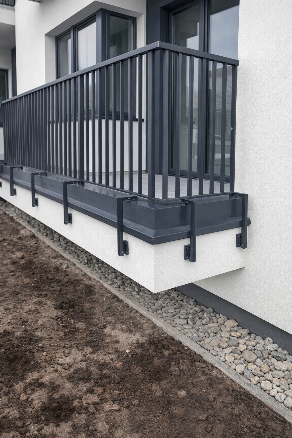 Nowoczesna, grafitowa balustrada balkonowa z pionowymi szczeblami, zamocowana na białej, betonowej podstawie. Widok z dołu, z fragmentem ziemi i kamieni ozdobnych.