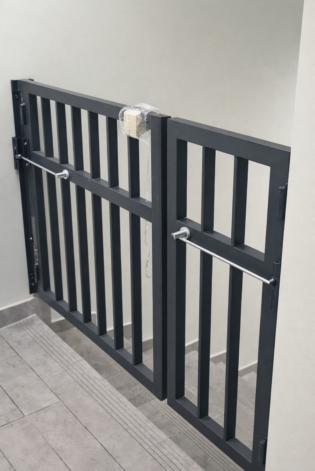 Metalowa barierka schodowa w kolorze grafitowym, z poziomymi szczeblami i zabezpieczeniem na środku, zamontowana na schodach z szarych płytek. Widoczne zawiasy i elementy mocujące.