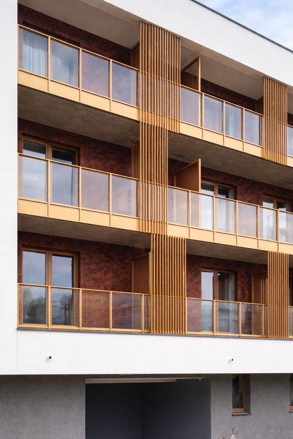 Nowoczesny budynek z balkonami z balustradami szklanymi i drewnianymi elementami dekoracyjnymi. Ujęcie z dołu, podkreślające geometrię i detale wykończenia.