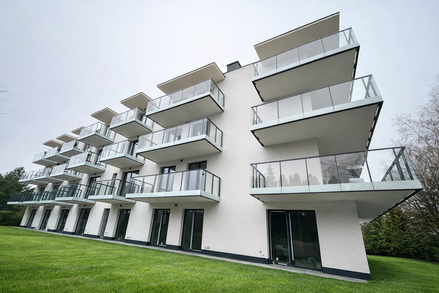 Nowoczesny budynek z białymi balkonami i szklanymi balustradami, widok z dołu na tle zielonego trawnika i pochmurnego nieba. Minimalistyczna architektura.