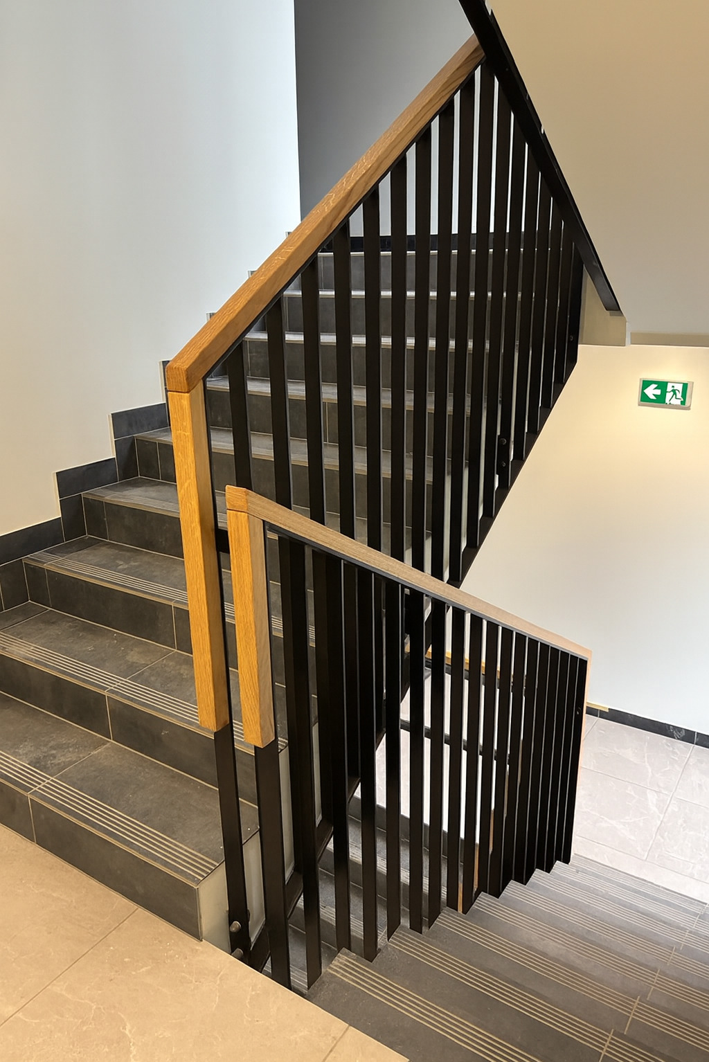 Nowoczesne schody z czarną metalową balustradą i drewnianymi poręczami, widok z góry. Szare płytki na schodach z delikatnym wzorem. Na ścianie znak ewakuacyjny.