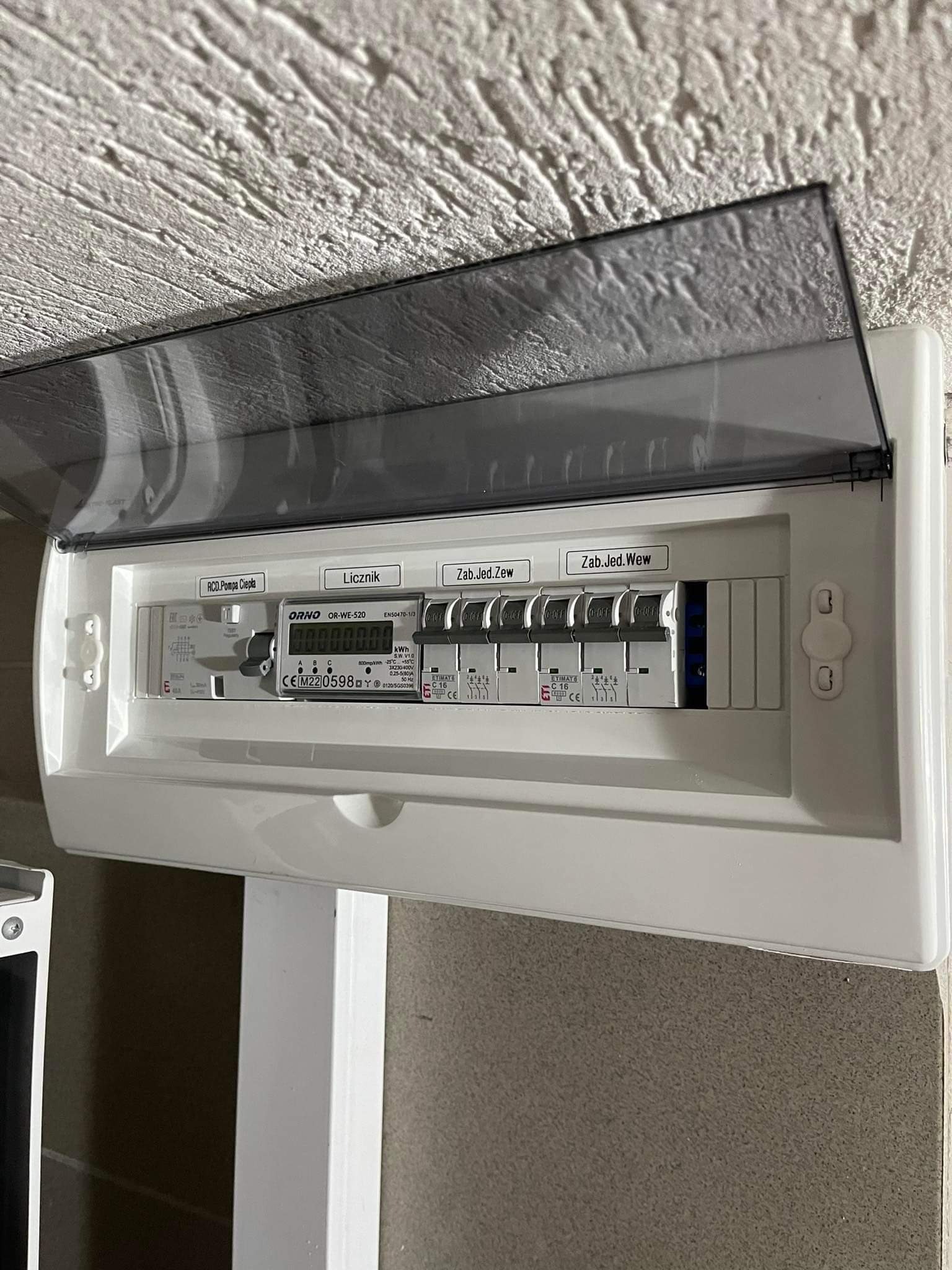 Rozdzielnica elektryczna ORNO z licznikiem i bezpiecznikami, zamontowana na ścianie o nierównej fakturze, z etykietami 'RCD.Pompa Ciepła', 'Licznik', 'Zab.Jed.Zew', 'Zab.Jed.Wew' i częściowo...