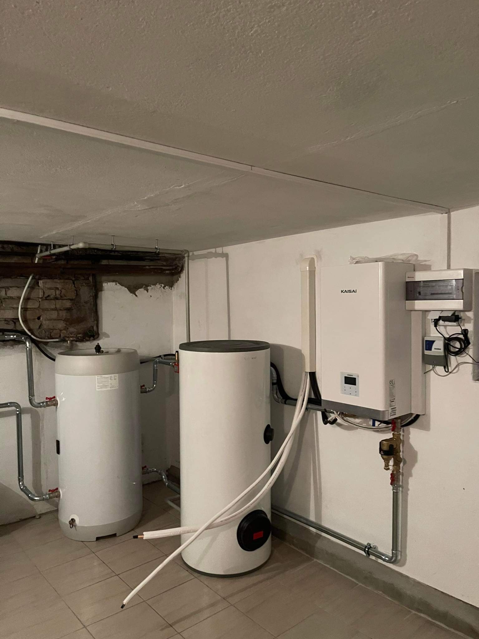 Dwa białe zasobniki ciepłej wody użytkowej oraz pompa ciepła marki Kaisai zamontowane w piwnicy z widocznymi instalacjami hydraulicznymi i elektrycznymi.