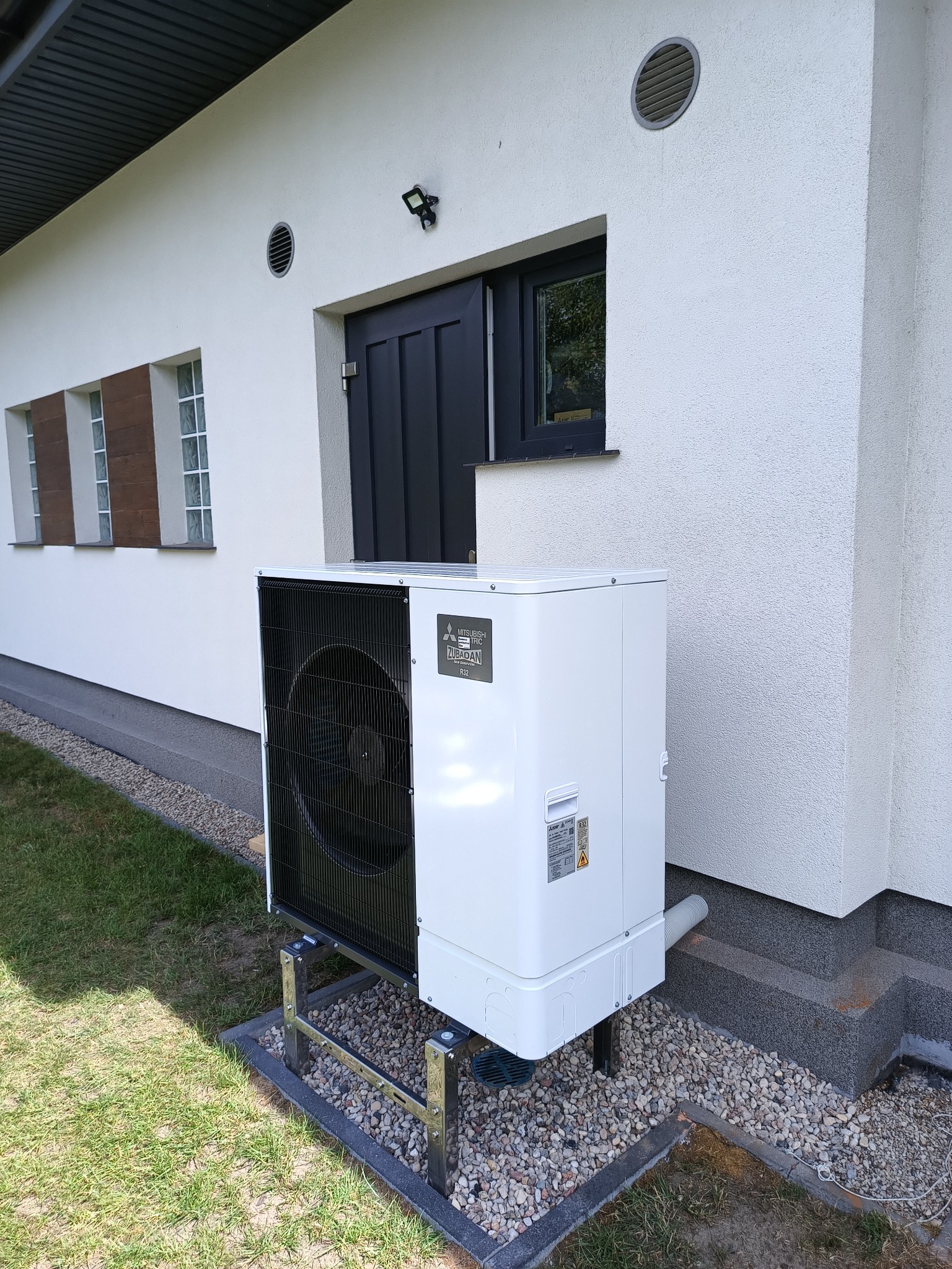 Biała pompa ciepła Mitsubishi Zubadan, zamontowana na metalowej konstrukcji, stoi na żwirowym podłożu przy białej ścianie domu z szarymi detalami.