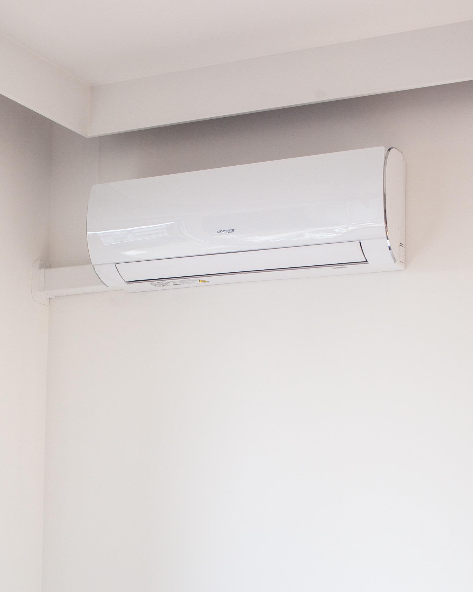 Biały klimatyzator marki Sinclair zamontowany na białej ścianie w rogu pomieszczenia, widoczne mocowanie urządzenia.