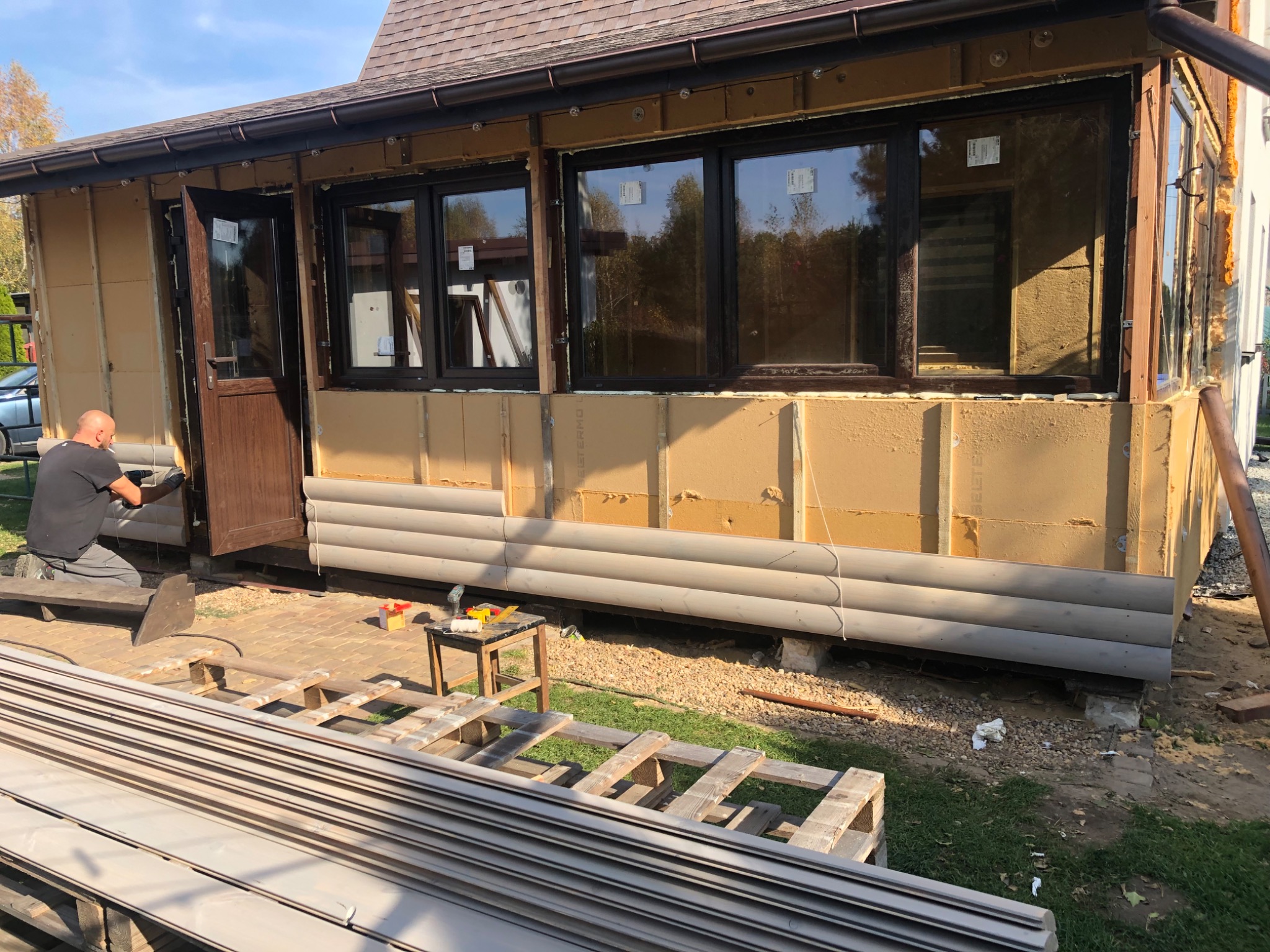 Prace przy ocieplaniu ścian zewnętrznych budynku z widoczną warstwą izolacyjną i drewnianą okładziną, pracownik montuje elementy wykończeniowe przy otwartych drzwiach wejściowych, na pierwszym...