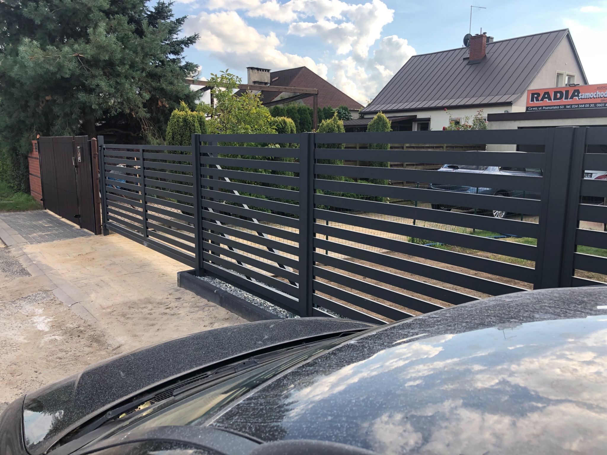 Nowoczesne, poziome, szare ogrodzenie panelowe z metalu, widok zza brudnej maski samochodu, w tle budynki mieszkalne i zieleń.