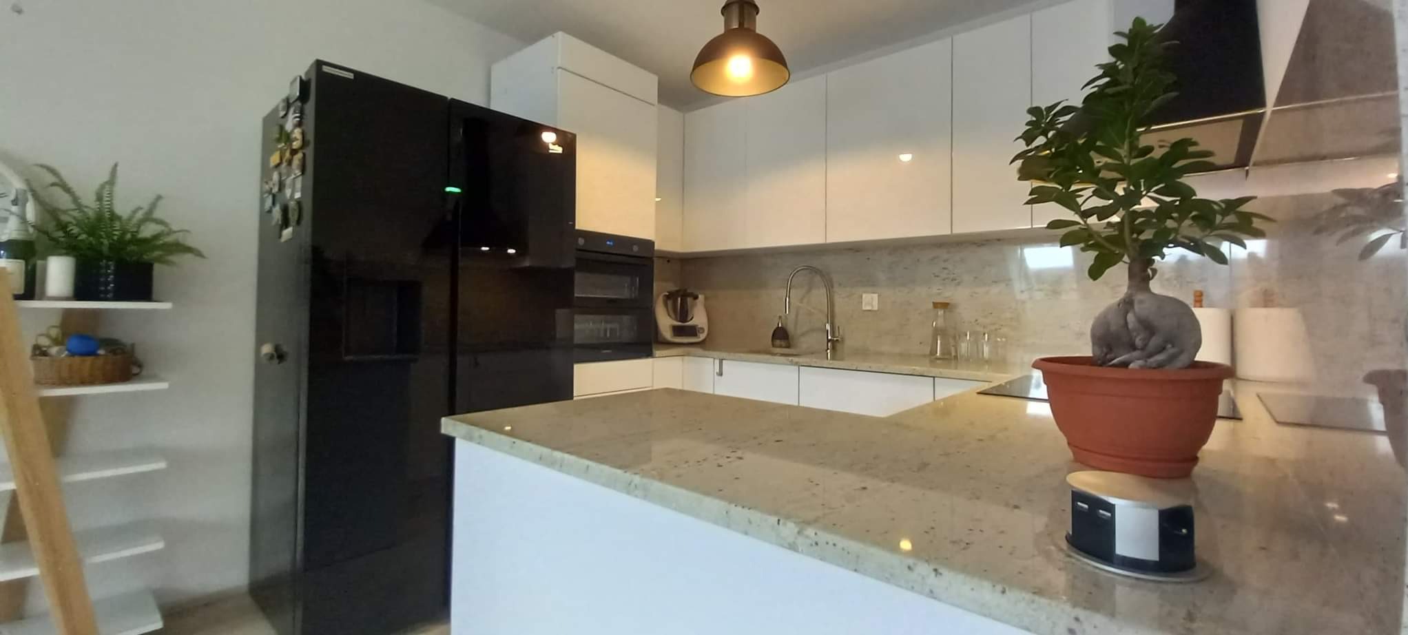 Jasny blat kamienny z otworem na wysuwaną listwę z gniazdami w nowoczesnej kuchni z czarną lodówką i białymi szafkami, oświetlony wiszącą lampą.