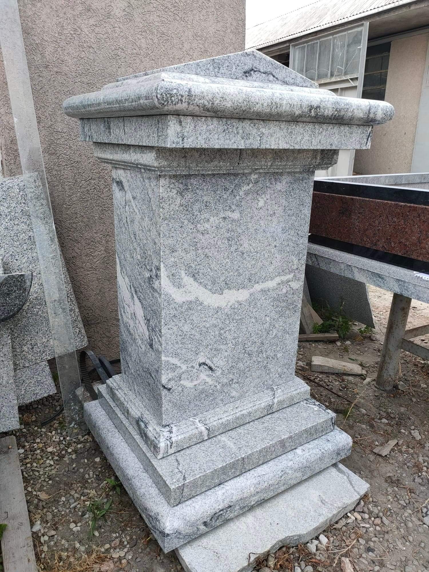 Gotowy, surowy, wysoki, granitowy postument z kwadratową podstawą i nakryciem, ustawiony na żwirowym podłożu w warsztacie kamieniarskim.