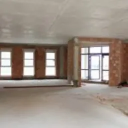 Wnętrze hali w trakcie budowy: surowy betonowy strop, ściany z czerwonej cegły dziurawki, trzy okna w jednej ścianie, otwór drzwiowy w innej, podłoże wylewka betonowa, widoczne narzędzia...