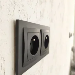 Dwa czarne gniazdka elektryczne z bolcem uziemiającym, zamontowane na ścianie o szorstkiej, jasnej fakturze, sfotografowane pod kątem.