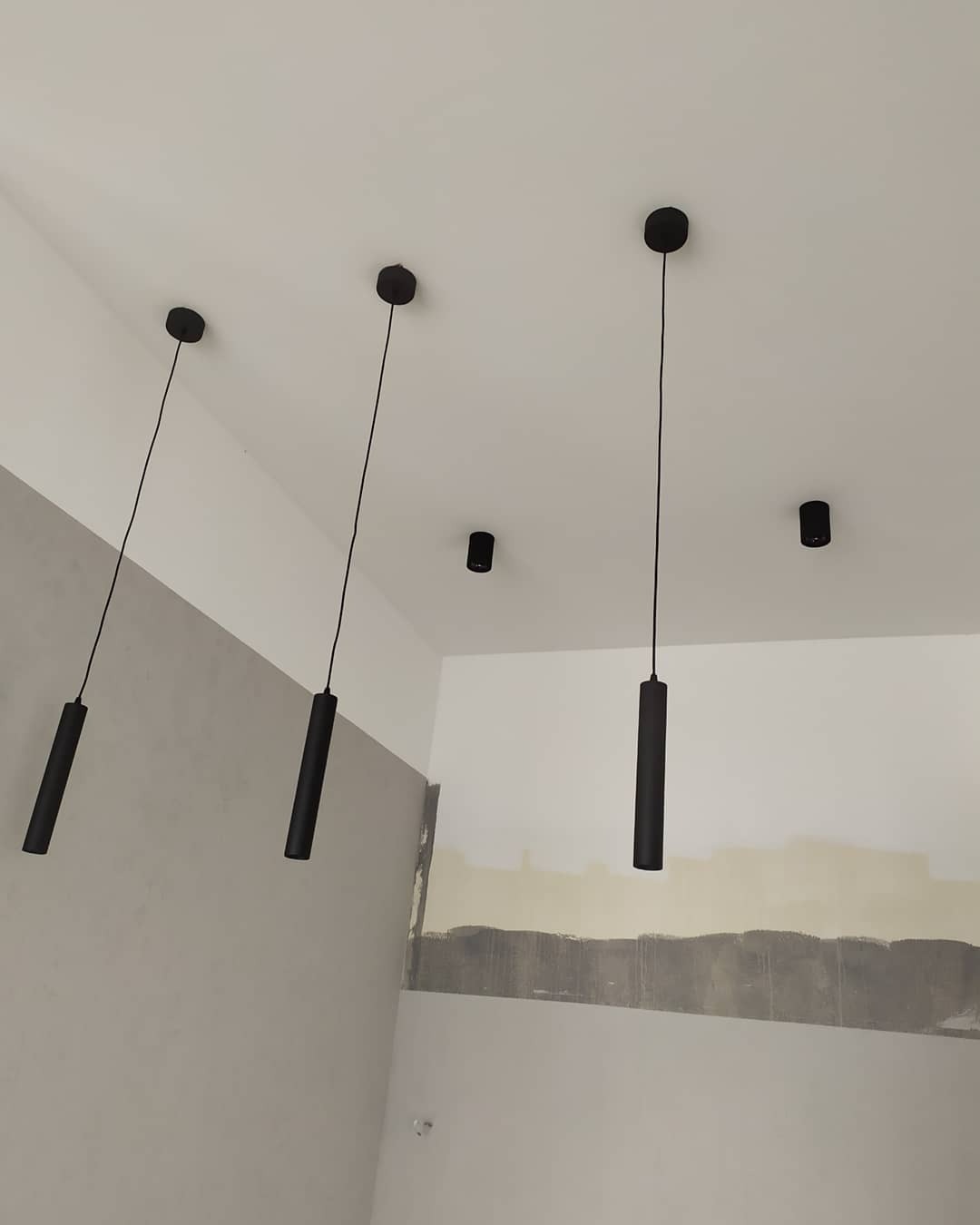 Cztery czarne lampy cylindryczne wiszące na cienkich kablach z sufitu w pomieszczeniu w trakcie remontu, widoczne ślady po malowaniu na ścianach.