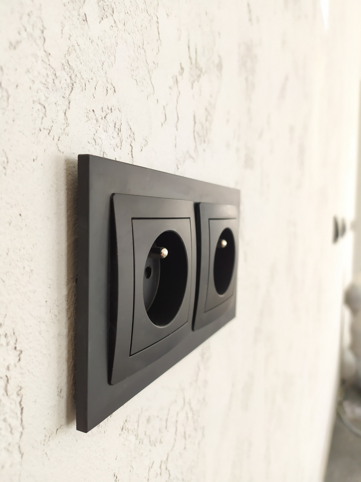 Dwa czarne gniazdka elektryczne z bolcem uziemiającym, zamontowane na ścianie o szorstkiej, jasnej fakturze, sfotografowane pod kątem.
