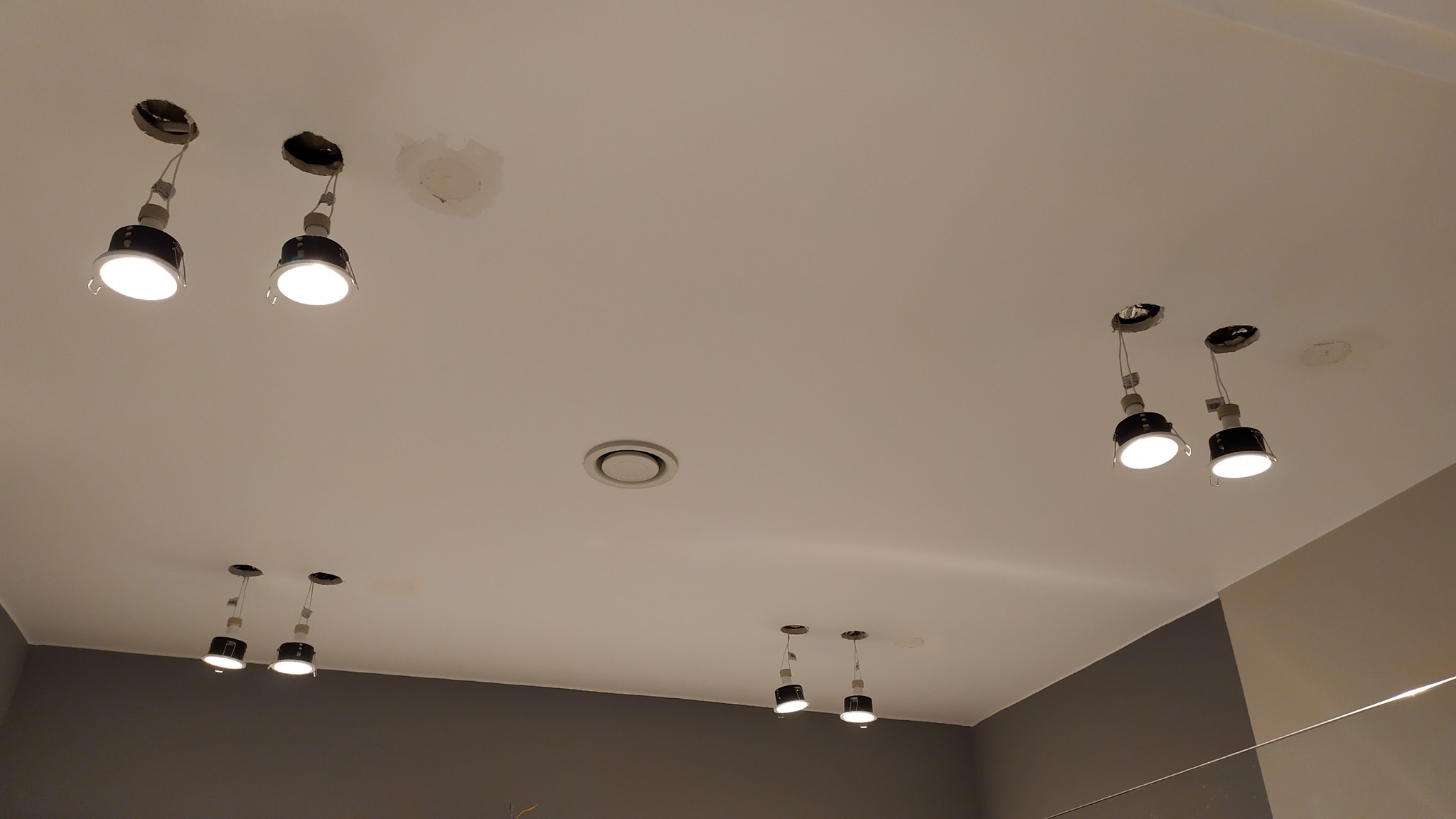 Biały sufit z zamontowanymi, ale jeszcze nie w pełni osadzonymi reflektorami punktowymi; widoczne przewody i otwory montażowe.