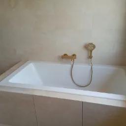 Biała wanna obudowana beżowymi płytkami imitującymi kamień, z zamontowaną złotą baterią i słuchawką prysznicową w nowoczesnej łazience.