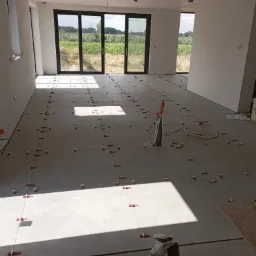 Przygotowanie do układania płytek podłogowych w przestronnym, jasnym pomieszczeniu z widokiem na zewnątrz przez duże okna; widoczne krzyżyki dystansowe i poziomica.