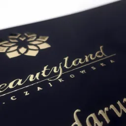 Elegancka wizytówka z czarnego papieru z tłoczonym złotym logo i tekstem 'Beautyland M. Czajkowska', w dolnym rogu widoczny fragment napisu 'darunkowy'.