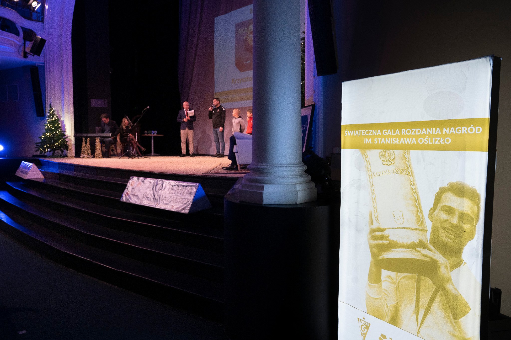 Podświetlany rollup prezentujący plakat 'Świąteczna Gala Rozdania Nagród im. Stanisława Oślizło' na tle sceny z występem artystycznym i choinką.
