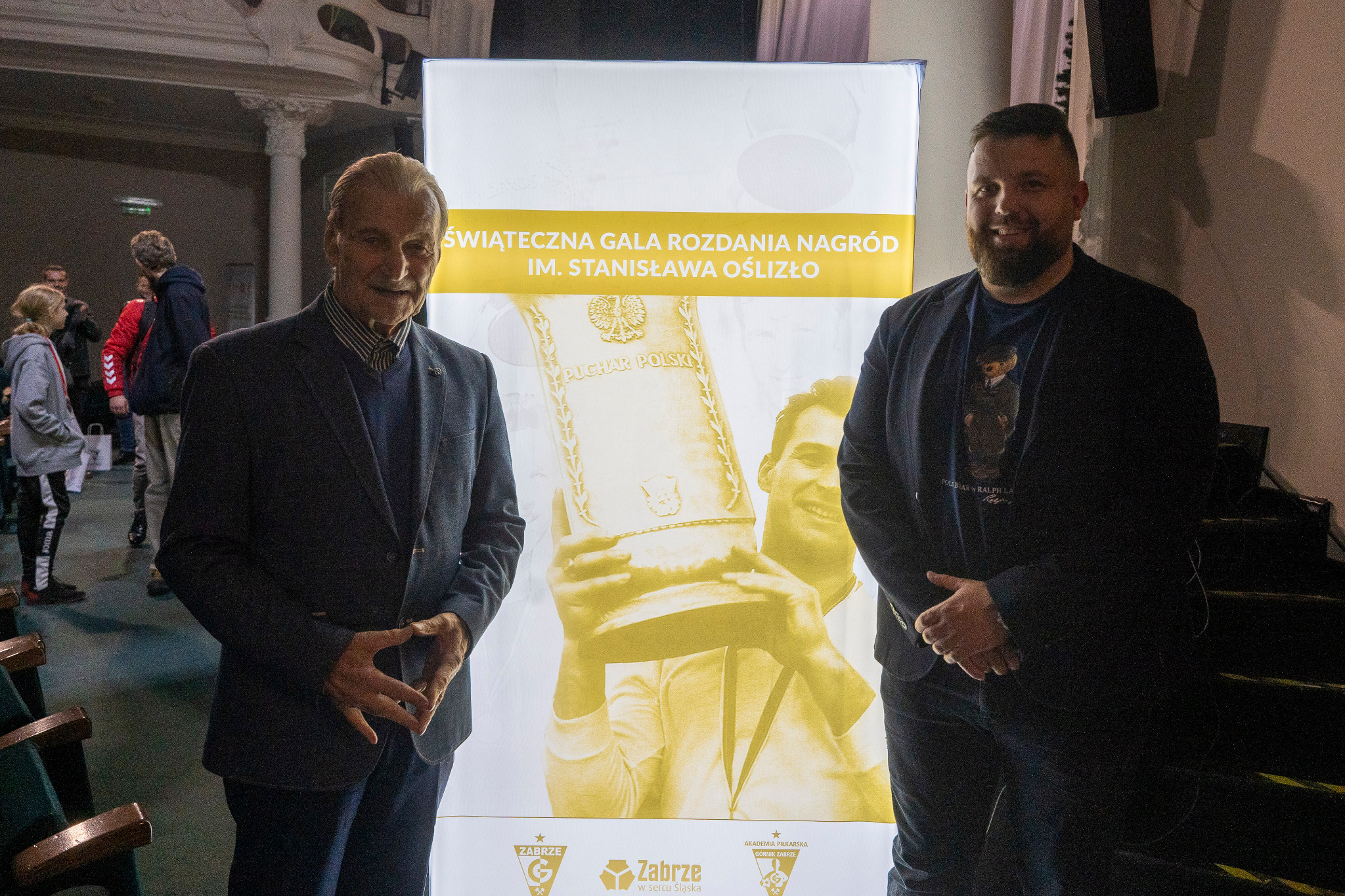 Dwaj mężczyźni pozują na tle rollupu z napisem 'Świąteczna Gala Rozdania Nagród im. Stanisława Oślizło' i grafiką Pucharu Polski. Widoczni także logotypy Zabrze i Akademii Piłkarskiej Górnik Zabrze.