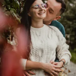 Uśmiechnięta dziewczyna w okularach i swetrze, obejmowana przez mężczyznę, który ją całuje w policzek na tle zielonych liści i czerwonych gałęzi.