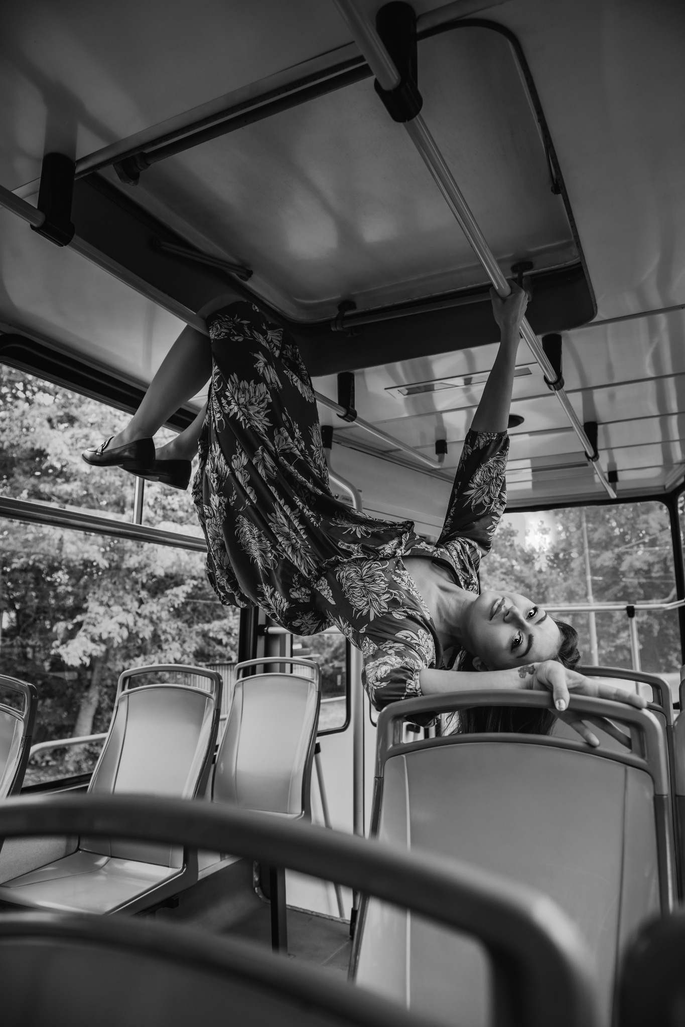 Kobieta w kwiecistej sukience, zwisająca głową w dół z poręczy w pustym tramwaju, czarno-biała fotografia z nietypowej perspektywy.