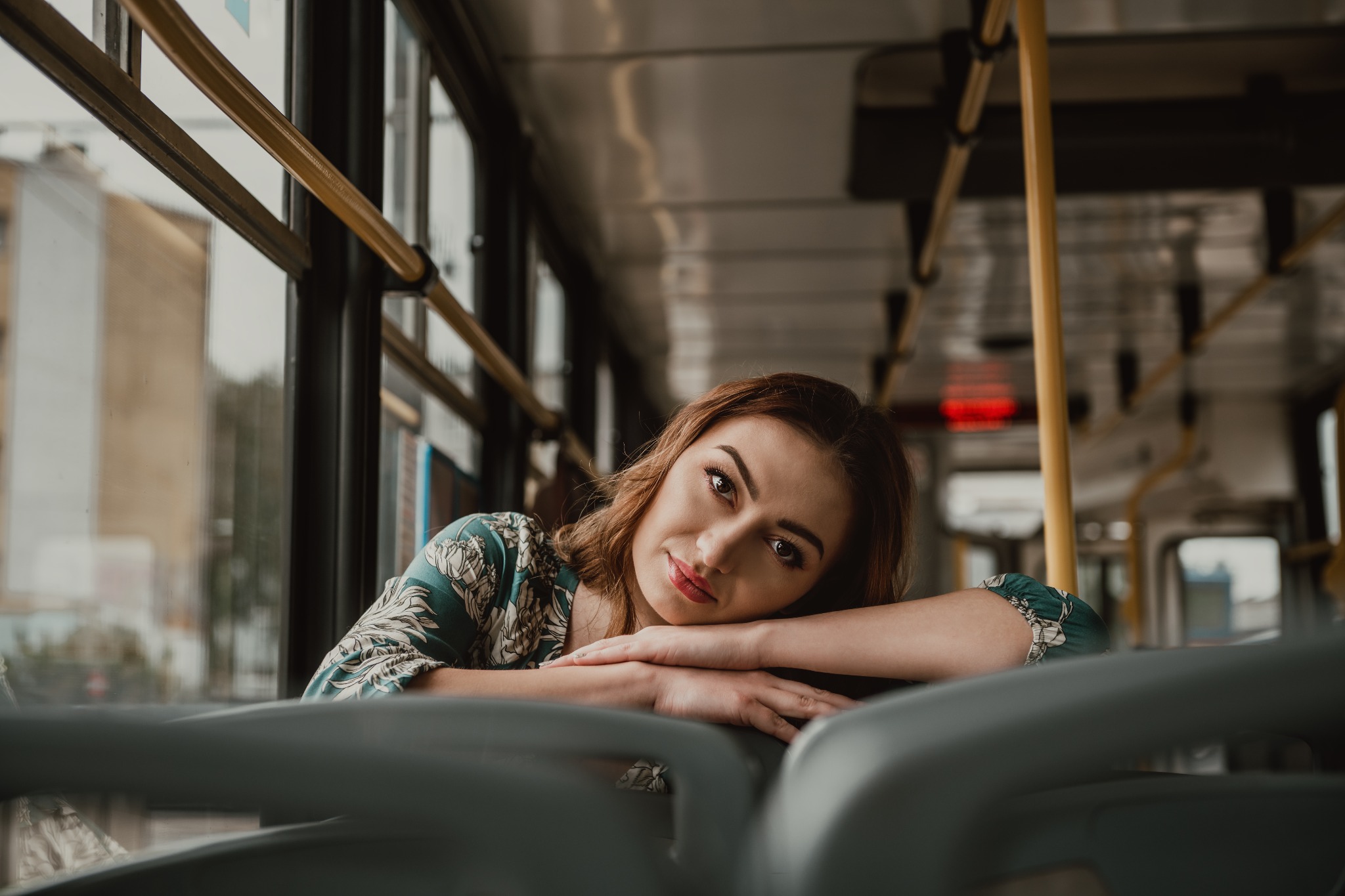 Kobieta w bluzce z motywem roślinnym opiera się na siedzeniu w autobusie, patrząc w obiektyw z lekkim uśmiechem.
