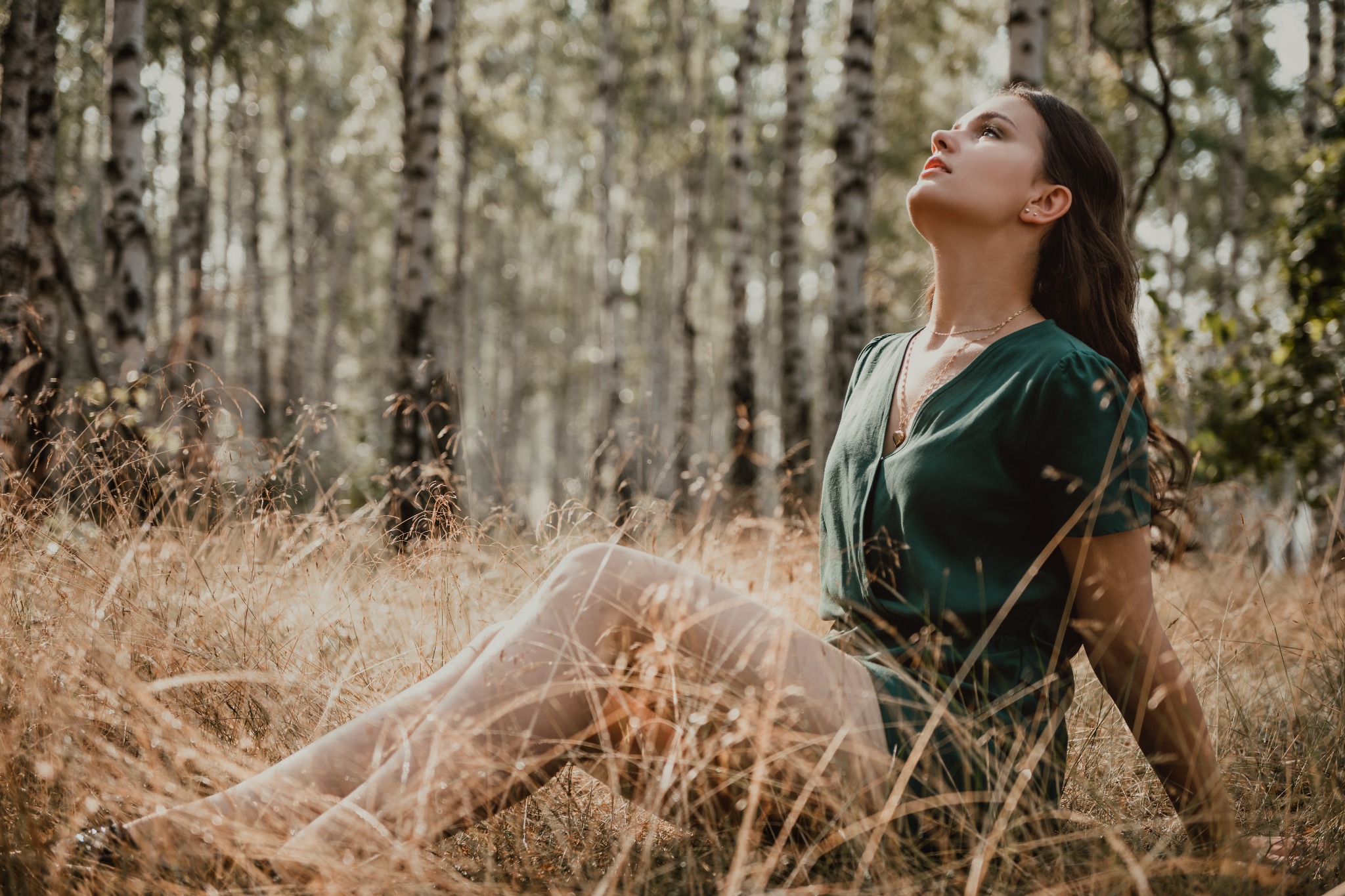 Młoda kobieta w zielonej sukience siedzi na trawie w lesie brzozowym, spoglądając w górę.