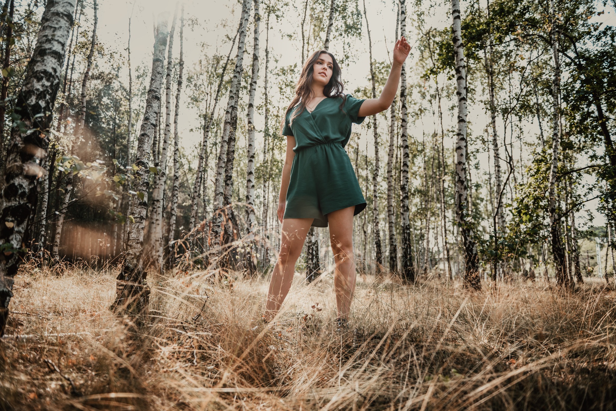 Młoda kobieta w zielonym kombinezonie pozuje w lesie brzozowym, z uniesioną ręką i delikatnym uśmiechem, w ciepłym świetle słonecznym.