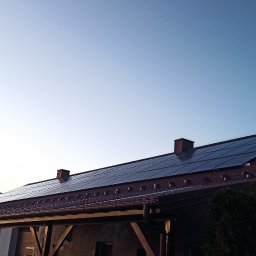 MAC-DAB - Panele słoneczne na dachu domu z brązową dachówką, widoczne kominy i antena telewizyjna, na pierwszym planie fragment samochodu dostawczego z bagażnikiem dachowym i drabina.