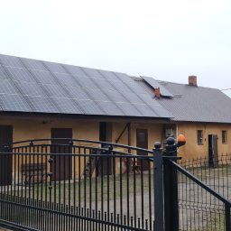 MAC-DAB - Instalacja paneli słonecznych na dachu budynku gospodarczego z żółtą elewacją, widok zza czarnego, metalowego ogrodzenia.