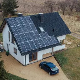 Widok z góry na biały dom jednorodzinny z ciemnym dachem pokrytym panelami słonecznymi; na podjeździe stoi niebieski samochód osobowy.